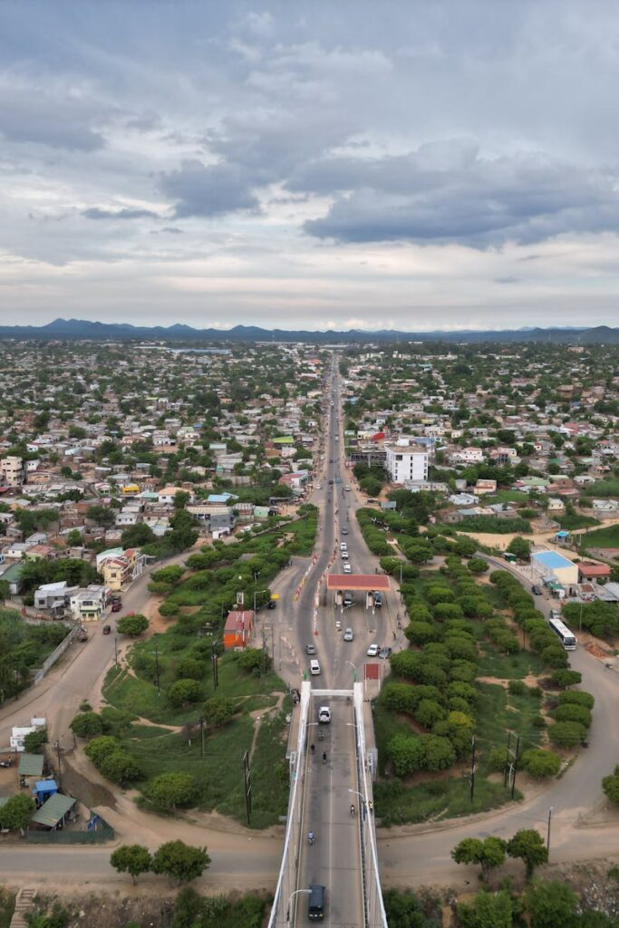 Wide aerial view of cityscape with central highway leading into the distance under a cloudy sky.